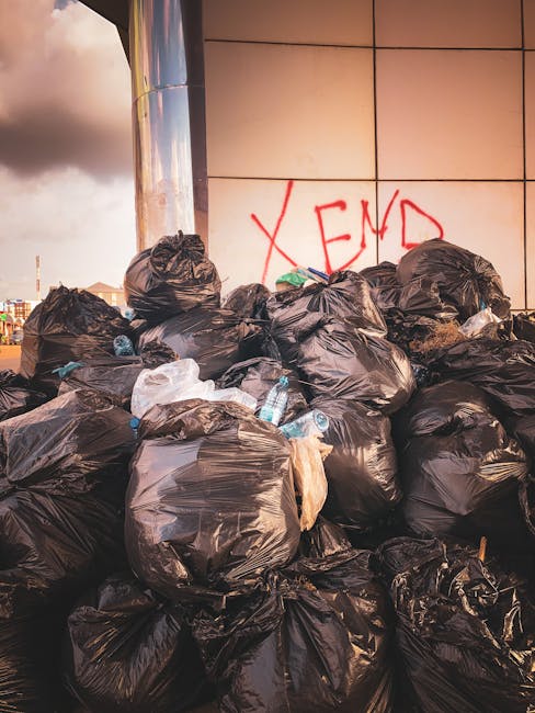 A large pile of black trash bags overflowing in front of a modern tiled wall with the word 'XEND' spray-painted in red graffiti. The bags appear tightly packed, some with visible plastic bottles and paper waste protruding. The scene is outdoors, likely on a pavement or driveway near an urban building, with the sky partly cloudy visible in the background. The lighting suggests late afternoon or early evening, creating soft shadows and a warm tone. This image illustrates a typical example of unmanaged waste accumulation, highlighting the importance of professional rubbish removal services like those offered by Rubbish Collection Catford for efficient on-site clearance and alternative waste handling options, especially in urban environments where clutter can be a concern.