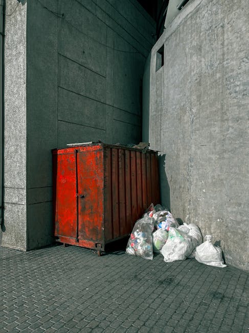 Four large black plastic garbage bags filled with waste material are positioned on a paved sidewalk near a wooden fence and a hedge with dense leafy branches. The bags appear tightly packed and are stacked against the fence, with some leaning slightly. The scene is outdoors, in what seems to be a residential or urban area, with a road running parallel to the sidewalk in the foreground. Ambient lighting suggests an overcast day or low-light conditions, creating a subdued, shadowed atmosphere. The presence of these waste bags indicates an on-site rubbish collection or private waste disposal service, as the bags are not in a typical refuse bin or skip. The environment appears clean apart from the bags, and there is no visible action occurring in the scene. This setup aligns with independent rubbish removal efforts, such as those provided by companies like Rubbish Collection Catford, operating in the local area and offering alternative waste handling solutions from standard council services.