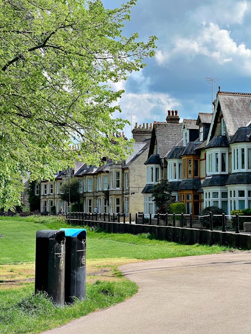 A public park area with lush green grass and a large leafy tree on the left side of the image. In the foreground, there are two black waste bins positioned on a paved pathway, one slightly taller and wider than the other. Behind the bins, a narrow strip of grass separates the pathway from a black wooden picket fence that borders a row of terraced Victorian-style houses with painted stone facades, bay windows, and tiled roofs, some with chimneys protruding. The houses are set on a slightly elevated position, with small front gardens and greenery visible between their facades. The sky above shows partly cloudy weather with patches of blue, creating a bright, natural daylight atmosphere. The scene is quiet and residential, with visual elements suggesting that the area might be serviced by independent rubbish collection or private waste handling, as indicated by the presence of the waste bins near the houses during daytime.