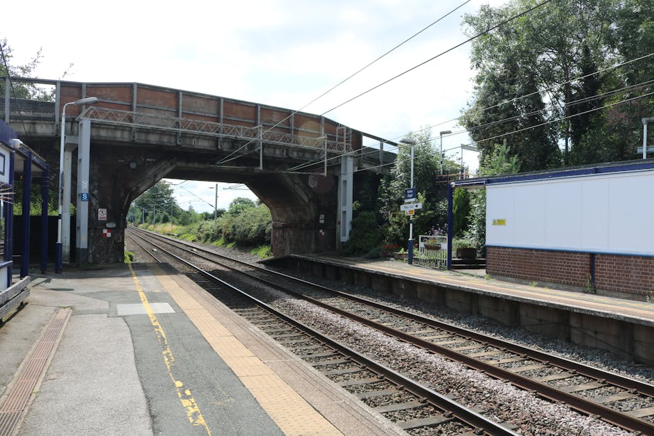 The image shows a railway station platform with train tracks running parallel to the edge. In the background, an arched brick and concrete bridge spans over the tracks, displaying weathered surfaces and a mix of reddish-brown bricks and grey concrete. To the left, there is a small shelter with blue metal framing and a translucent roof, positioned just behind the yellow tactile paving that runs along the platform edge, providing accessible guidance for visually impaired passengers. On the right side, a white brick building with a yellow warning sign is visible, with a small bench or seating area near it. Overhead, electrical wires stretch across the scene, supported by poles, indicative of electric train services. Surrounding greenery and trees are seen beyond the station, adding a natural backdrop to the urban infrastructure. The lighting suggests a partly cloudy day, with diffuse sunlight illuminating the scene evenly, typical of a peaceful, non-rush hour setting. Such a station and its surrounding structures may be relevant to independent waste disposal and rubbish collection services that support station maintenance and nearby residential or commercial waste management, such as Rubbish Collection Catford.