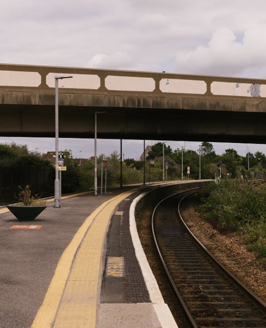 The image shows a railway station platform with train tracks running parallel to the edge. In the background, an arched brick and concrete bridge spans over the tracks, displaying weathered surfaces and a mix of reddish-brown bricks and grey concrete. To the left, there is a small shelter with blue metal framing and a translucent roof, positioned just behind the yellow tactile paving that runs along the platform edge, providing accessible guidance for visually impaired passengers. On the right side, a white brick building with a yellow warning sign is visible, with a small bench or seating area near it. Overhead, electrical wires stretch across the scene, supported by poles, indicative of electric train services. Surrounding greenery and trees are seen beyond the station, adding a natural backdrop to the urban infrastructure. The lighting suggests a partly cloudy day, with diffuse sunlight illuminating the scene evenly, typical of a peaceful, non-rush hour setting. Such a station and its surrounding structures may be relevant to independent waste disposal and rubbish collection services that support station maintenance and nearby residential or commercial waste management, such as Rubbish Collection Catford.