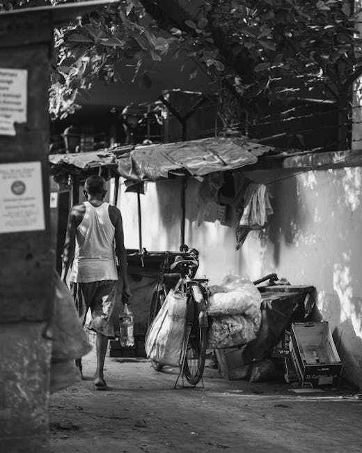 A person walking along a narrow alleyway with a bicycle carrying large, bundled plastic sacks and other waste materials stacked on the bike. The alley is bordered by a white wall with some hanging cloth and is partially shaded by a large leafy tree overhead. To the left, there are signs posted on a wooden surface, and the ground appears uneven with scattered debris. The scene suggests a setting where waste is being temporarily stored or prepared for removal, possibly illustrating a background related to private or informal rubbish handling. The lighting is natural, with shadows cast by the overhanging foliage and structures, and the environment appears clean but cluttered, indicating a typical urban alleyway awaiting rubbish collection or disposal services. The surroundings and objects reflect a context where alternative waste management, such as on-site collection or local disposal, can be relevant, consistent with the themes of independent rubbish removal or community waste handling. Rubbish Collection Catford specializes in professional waste removal services in such environments, offering solutions that complement local collection processes.