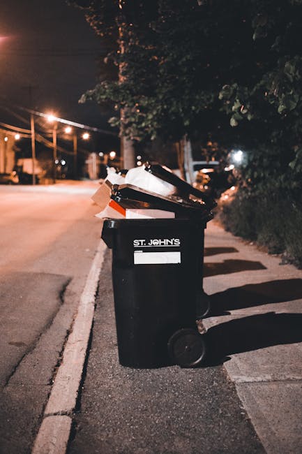 A person walking along a narrow alleyway with a bicycle carrying large, bundled plastic sacks and other waste materials stacked on the bike. The alley is bordered by a white wall with some hanging cloth and is partially shaded by a large leafy tree overhead. To the left, there are signs posted on a wooden surface, and the ground appears uneven with scattered debris. The scene suggests a setting where waste is being temporarily stored or prepared for removal, possibly illustrating a background related to private or informal rubbish handling. The lighting is natural, with shadows cast by the overhanging foliage and structures, and the environment appears clean but cluttered, indicating a typical urban alleyway awaiting rubbish collection or disposal services. The surroundings and objects reflect a context where alternative waste management, such as on-site collection or local disposal, can be relevant, consistent with the themes of independent rubbish removal or community waste handling. Rubbish Collection Catford specializes in professional waste removal services in such environments, offering solutions that complement local collection processes.