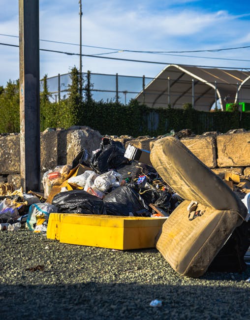 A pile of mixed waste materials and rubbish bags collected outside on a gravel surface next to a low stone wall, with a yellow container or tray lying among the debris. The waste includes black and transparent plastic bags, some contents spilling out, and various discarded items such as an old, dirty beige upholstered car seat or cushion leaning against the rubbish for disposal. In the background, a tall metal utility pole with power lines, a hedge, and a sports facility with a curved, partially covered athletic field or tennis court are visible under a blue sky with some light clouds. The scene suggests an area prepared for private rubbish removal, with the debris awaiting collection by a waste management service like Rubbish Collection Catford. Natural sunlight illuminates the scene, highlighting the textures of the discarded materials and contrasting the tidy environment with the accumulated waste, illustrating a typical site for non-regulated, independent rubbish disposal or clearance in the context of alternative waste handling options.
