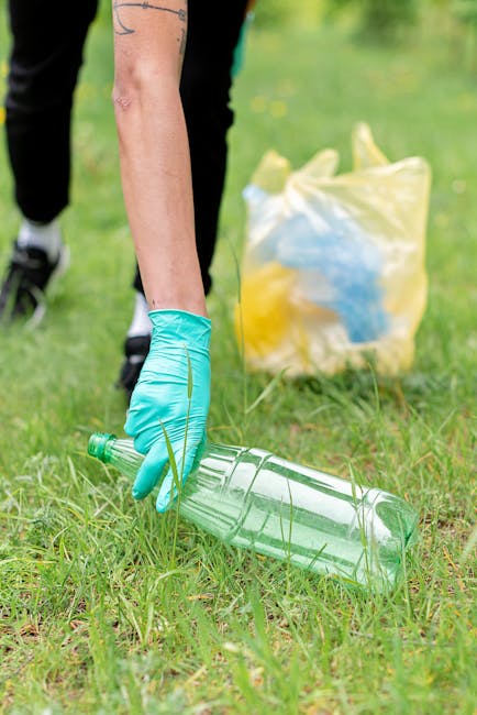 A person wearing a light blue rubber glove is seen picking up a clear plastic bottle from a grassy area, with a yellow and blue rubbish bag in the background. The individual’s arm is extended downward, and the bottle appears to be a typical PET beverage container with a greenish tint. The surrounding environment features green grass and small yellow flowers, indicative of an outdoor setting such as a park or garden. The scene is illuminated by natural daylight, highlighting the textures of the grass and the shiny surface of the plastic bottle. In the background, the bag appears to contain additional waste or recyclables, and the person's clothing, partially visible, includes dark pants and sneakers. This scene reflects local or private waste collection efforts, typical of initiatives by companies like Rubbish Collection Catford, emphasizing service for rubbish removal or on-site clearance in a natural environment.