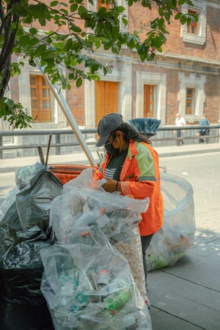 A pile of mixed waste materials and rubbish bags collected outside on a gravel surface next to a low stone wall, with a yellow container or tray lying among the debris. The waste includes black and transparent plastic bags, some contents spilling out, and various discarded items such as an old, dirty beige upholstered car seat or cushion leaning against the rubbish for disposal. In the background, a tall metal utility pole with power lines, a hedge, and a sports facility with a curved, partially covered athletic field or tennis court are visible under a blue sky with some light clouds. The scene suggests an area prepared for private rubbish removal, with the debris awaiting collection by a waste management service like Rubbish Collection Catford. Natural sunlight illuminates the scene, highlighting the textures of the discarded materials and contrasting the tidy environment with the accumulated waste, illustrating a typical site for non-regulated, independent rubbish disposal or clearance in the context of alternative waste handling options.